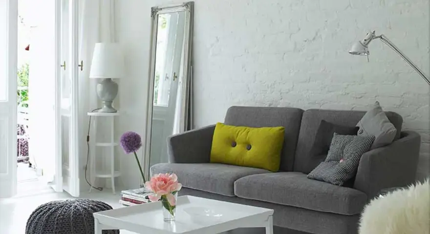 Sofá gris con cojines, espejo alto y lámpara. Trucos para decorar espacios pequeños con estilo y amplitud.