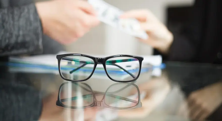 Okulary na pierwszym planie, w tle ręce przekazujące pieniądze. Idealny moment na skorzystanie z promocji na okulary!