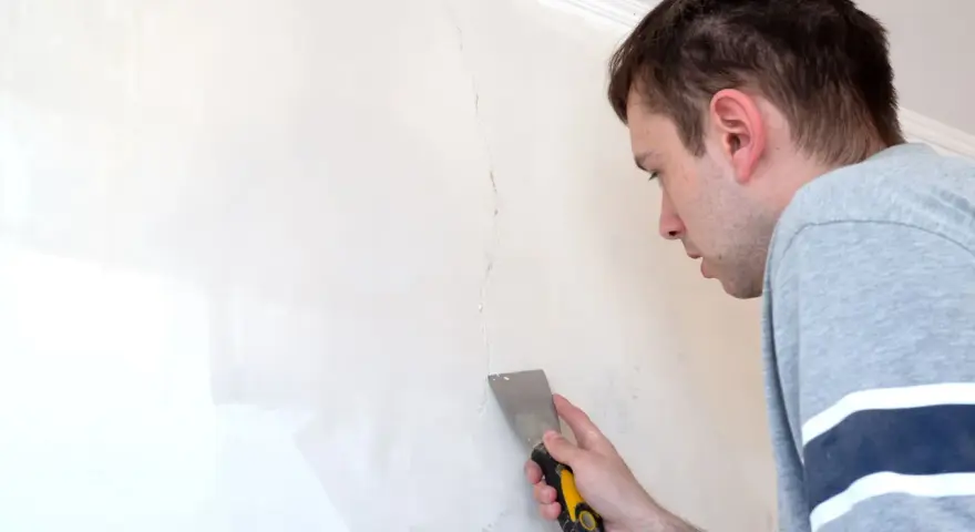 Uomo con spatola ripara una crepa su un muro bianco.