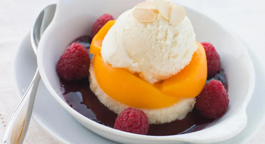 Ein köstlicher Pfirsich Melba: Vanilleeis auf Pfirsichspalten, umgeben von Himbeeren und dunkler Sauce, garniert mit Mandelblättchen.