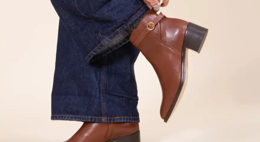 Une femme ajuste sa tenue avec bottines marron. Un jean évasé et un pull blanc complètent le look.