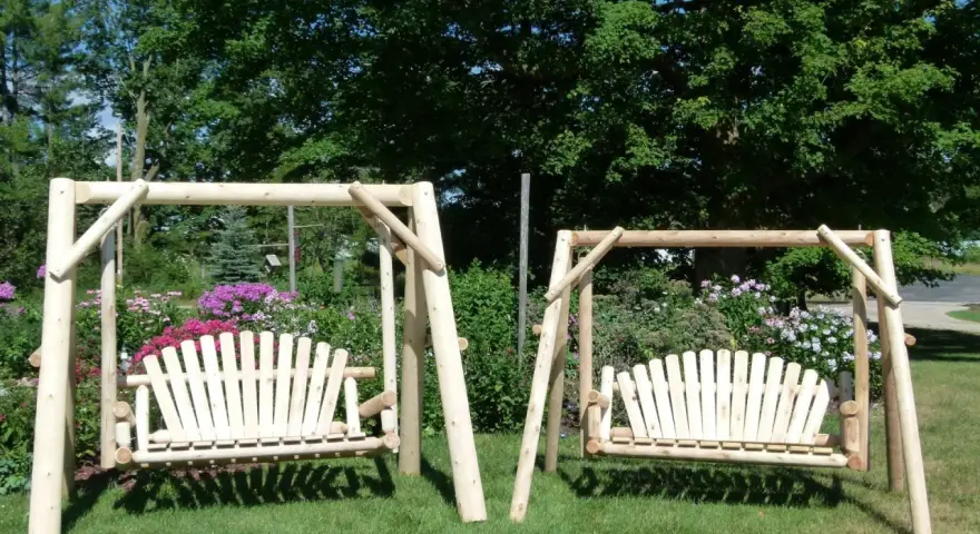 Two wooden porch swings stand on a grassy lawn, surrounded by colorful flowers and lush trees. This image offers inspiration for how to hang a porch swing.