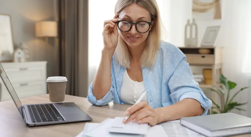 Femme aux lunettes calculant, laptop et café à proximité. Elle semble gérer des finances, peut-être une réduction de capital non motivée par des pertes.