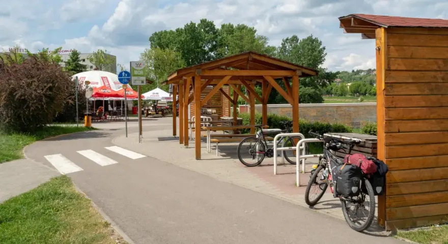 Miejsce odpoczynku przy ścieżce rowerowej. Dwa rowery, w tym jeden z sakwami, czekają na dalszą podróż. W tle zadaszenie i stoliki, idealne na przerwę w trasie.