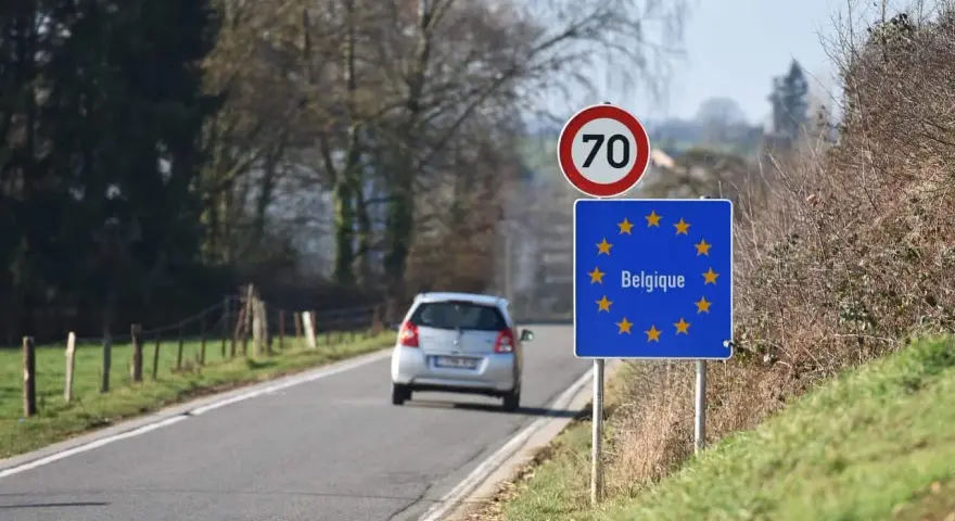 Znak graniczny Belgii, gdzie potrzebny paszport, na tle drogi i jadącego samochodu.