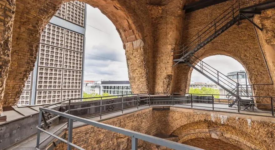 Blick durch die Bögen der Kaiser-Wilhelm-Gedächtniskirche auf eine moderne Glasfassade und Treppen.