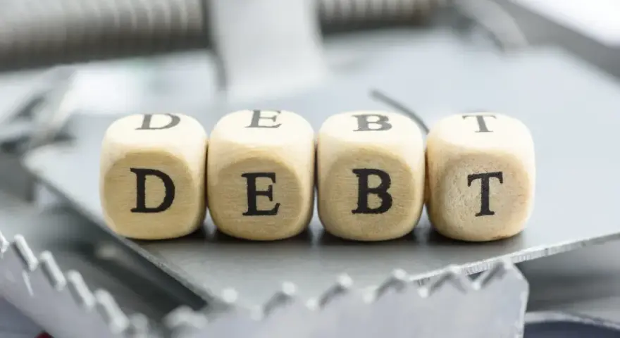 Cztery drewniane kostki z literami D, E, B, T tworzą słowo "DEBT" na pułapce. To symbol finansowych zobowiązań, które mogą prowadzić do trudnej sytuacji, jak datio in solutum.