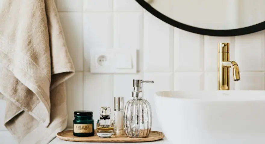 Sur un plateau en bois, des flacons de parfum et un distributeur de savon argenté. Une serviette beige est suspendue à côté d'un lavabo blanc et d'un robinet doré. La colle carrelage flex, c'est quoi ?