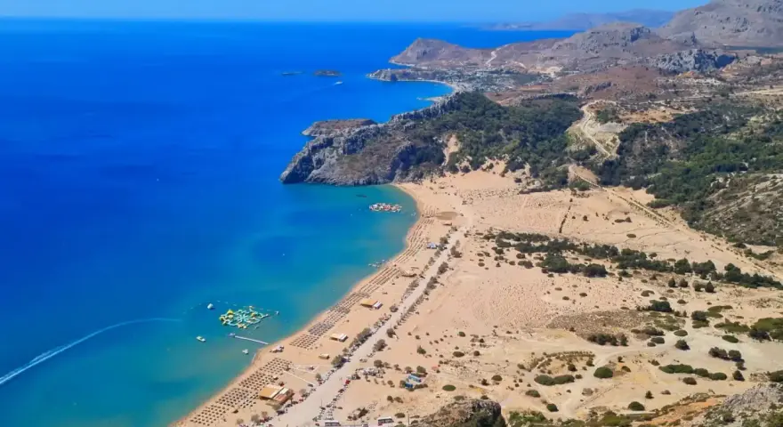 Malownicze **Rodos plaże** z turkusową wodą, piaszczystym brzegiem i zielonymi wzgórzami w tle. Widać wodny plac zabaw.