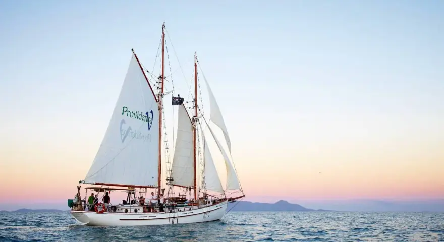 Żaglowiec "Providence" płynie po spokojnych wodach, zbliżając się do Whitehaven Beach. Zachód słońca maluje niebo pastelowymi barwami.