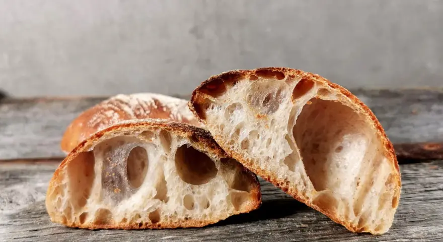 Zwei Hälften eines rustikalen ciabatta mit sauerteig, mit großer, luftiger Krume und knuspriger Kruste, liegen auf einem Holzbrett.