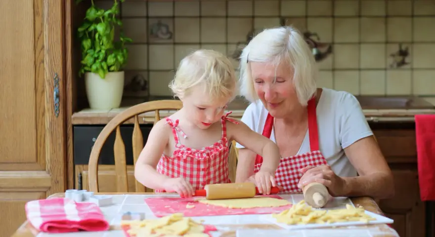 Babcia z wnuczką pieką ciasteczka. Dziewczynka wałkuje ciasto, a babcia z uśmiechem patrzy na jej pracę.