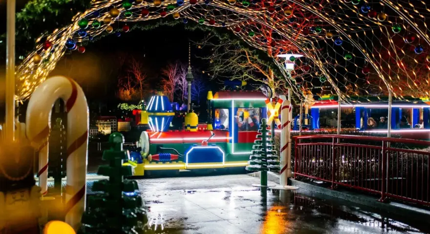 Ein bunter Zug fährt durch eine festlich beleuchtete Gasse im winterwonder legoland. Lichterketten und Kugeln schmücken den Weg.