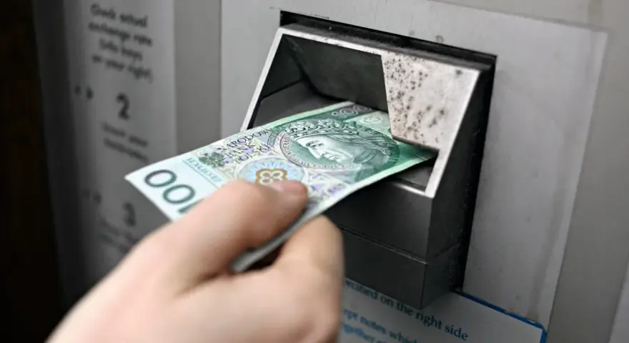 Ręka wkłada banknot 100 zł do bankomatu. Czy państwo może zabrać pieniądze z konta w razie wojny?