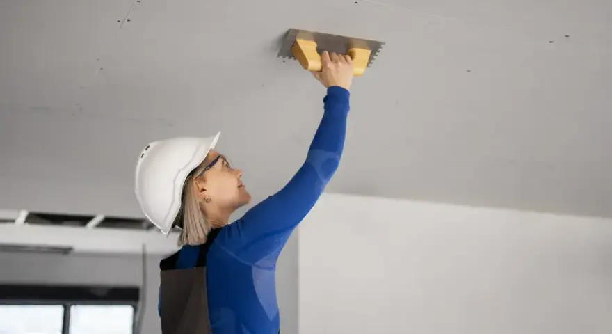 Kobieta w kasku i okularach wygładza tynk na suficie podwieszanym w łazience.