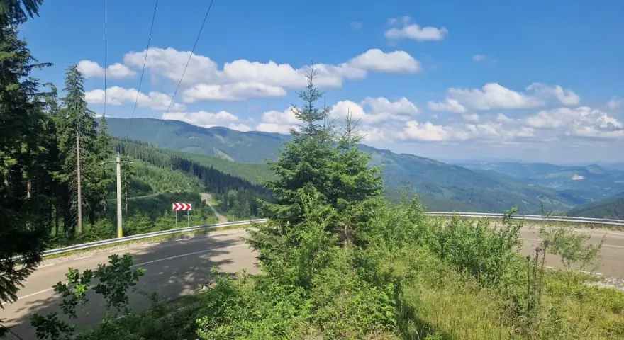 Górzysty krajobraz Rumunii z krętą drogą i lasami. W oddali widać pasma górskie, a na niebie chmury.