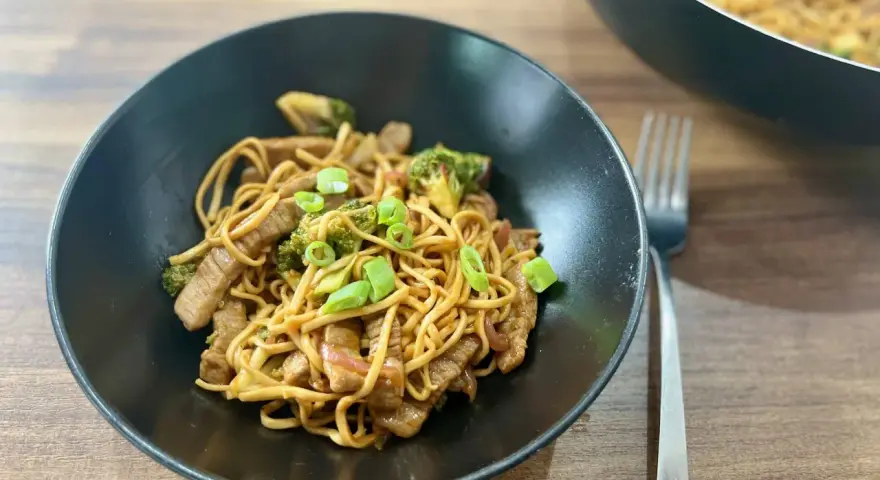 Pyszny makaron wok z kawałkami wołowiny i brokułami, posypany szczypiorkiem. Danie prezentuje się apetycznie w czarnej misce.