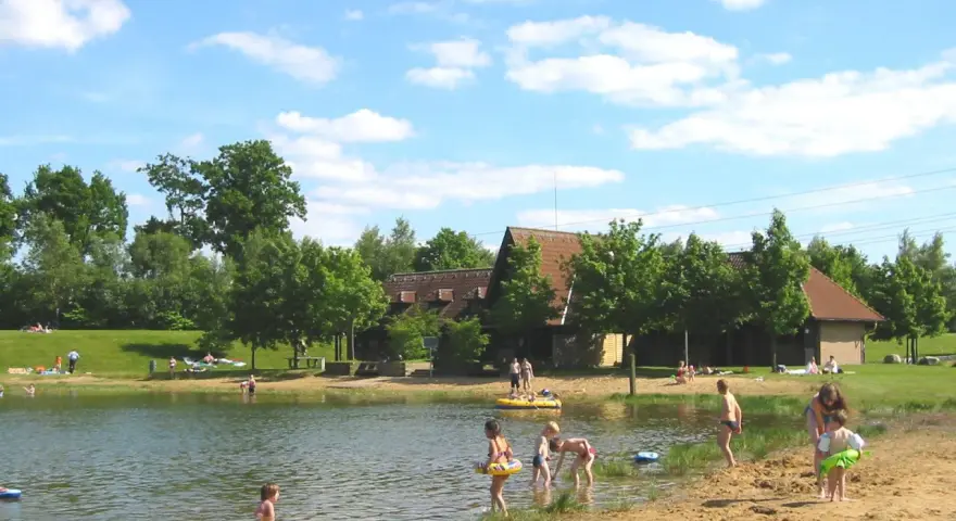 Sonniger Tag am badesee fredenbeck. Kinder planschen im Wasser und am Sandstrand, während im Hintergrund Gebäude und Bäume zu sehen sind.