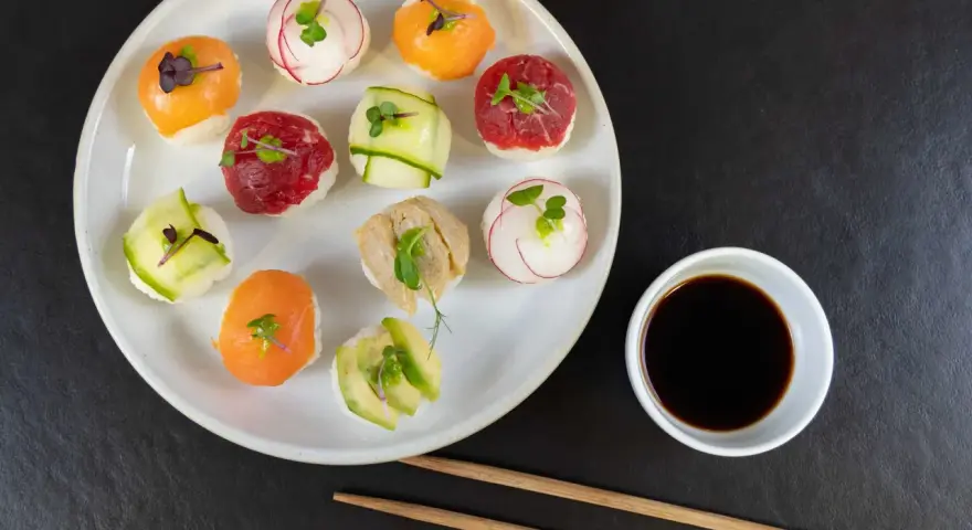 Na talerzu ułożone są kolorowe kulki sushi z łososiem, wołowiną, awokado i rzodkiewką. Obok miseczka sosu sojowego i pałeczki.