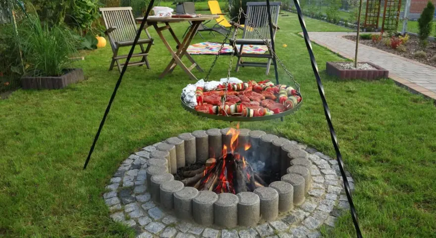 Ognisko w kamiennym kręgu, nad którym wisi okrągła taca z mięsem i warzywami na szaszłykach, gotowe do grillowania w ogrodzie.