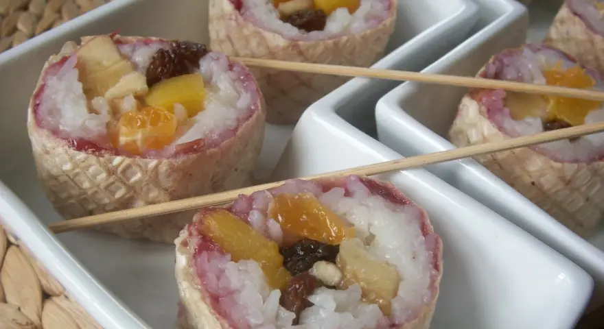 Słodkie sushi owocowe w wafelkach, z ryżem i kawałkami owoców, podane na białych tacach.