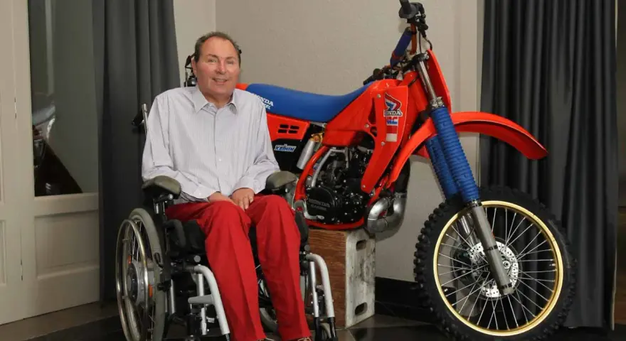 Andre Malherbe, leyenda del motocross, sonríe junto a su icónica moto Honda roja.