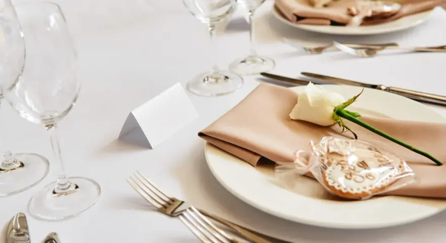 Elegancko nakryty stół weselny z różową serwetką, białą różą i pierniczkiem. Idealne miejsce na zastanowienie, jakie wino na wesele wybrać.