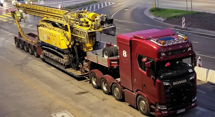 Czerwona ciężarówka Scania z przyczepą transportuje żółtą maszynę wiertniczą. Pilot pojazdu nienormatywnego czeka na przejazd.