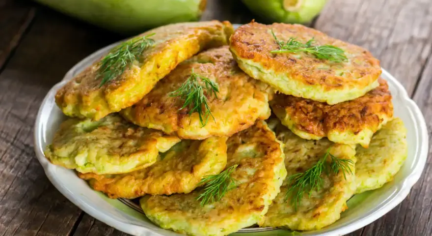 Pile de beignets de courgette dorés et moelleux, garnis de brins d'aneth frais, sur une assiette blanche. Deux courgettes vertes floues en arrière-plan.
