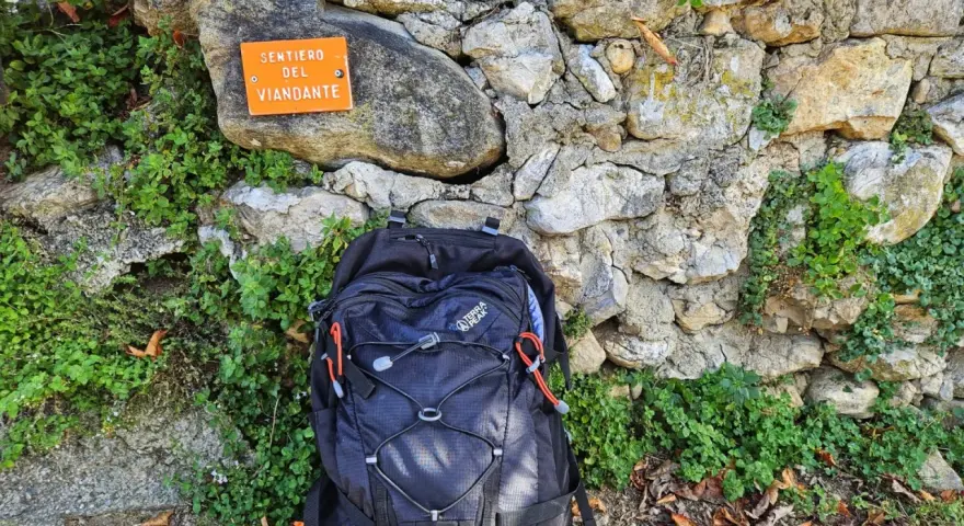 Zaino nero appoggiato a un muretto di pietra, accanto a un cartello arancione che indica "Sentiero del Viandante".