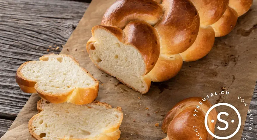 Ein goldbraunes Zopf Brot, frisch gebacken, liegt auf Backpapier. Zwei Scheiben sind abgetrennt und zeigen die weiche Krume.