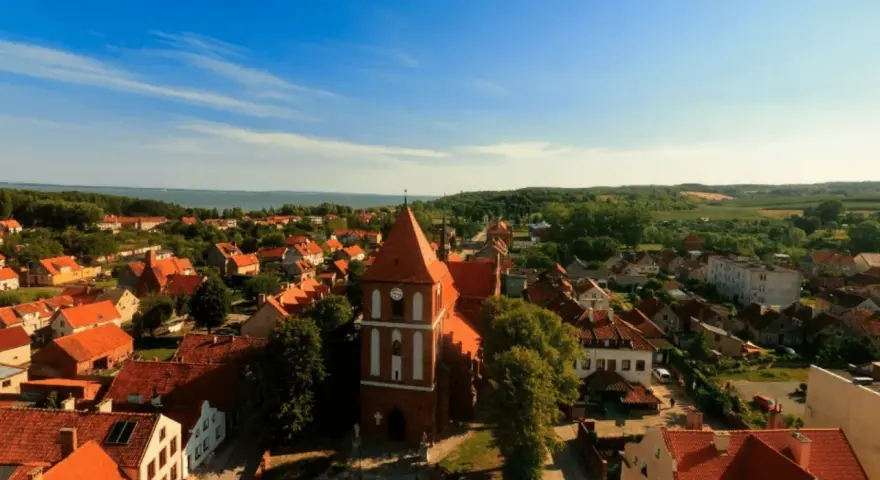 Widok na urokliwe Tolkmicko z charakterystyczną wieżą kościelną. Wśród czerwonych dachów i zieleni rozciąga się malowniczy krajobraz, idealny na odkrywanie tolkmickich atrakcji.