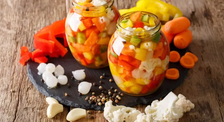 Due barattoli di giardiniera fatta in casa, pieni di verdure colorate come peperoni, carote e cavolfiore, su un tagliere di ardesia con ingredienti freschi intorno.
