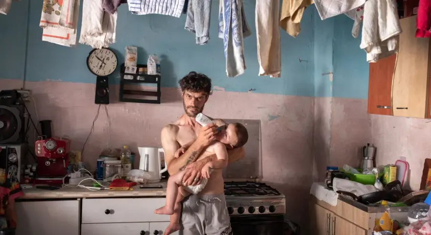 Emiliano Dante, padre single, nutre il suo bambino in una cucina disordinata, con panni stesi ad asciugare.