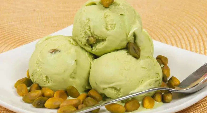 Tre palline di cremoso gelato al pistacchio, guarnite con pistacchi interi, su un piatto bianco con una spolverata di pistacchi tostati.