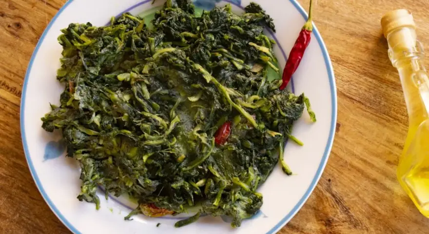 Un piatto di cicoria in padella, condita con peperoncino rosso e un filo d'olio d'oliva, su un tavolo di legno.