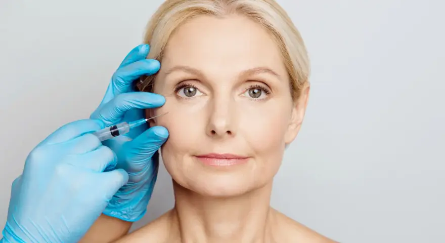 Zabieg z użyciem botoksu, czy botox jest bezpieczny? Dłoń w niebieskiej rękawiczce wykonuje zastrzyk w okolicę oka kobiety.