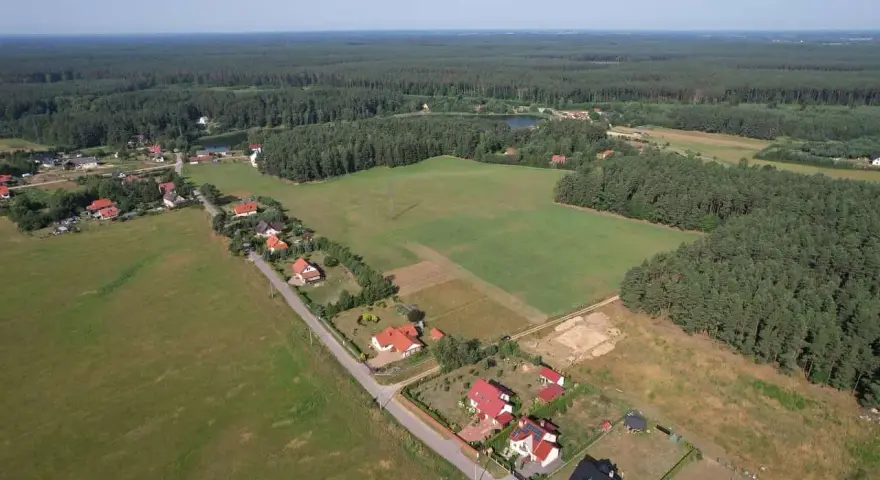 Widok z lotu ptaka na wieś otoczoną lasem. Widać domy z czerwonymi dachami, pola uprawne i jezioro. Każdy dom zajmuje swój kawałek ziemi, a całość tworzy malowniczy krajobraz.