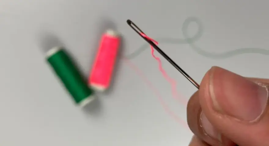 Dedos sujetan una aguja para meter hilos, con carretes de hilo verde y rosa desenfocados al fondo.