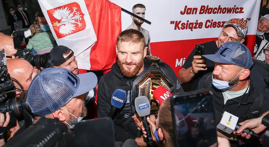 Jan Błachowicz, "Książę Cieszyński", mistrz UFC, otoczony przez fanów i dziennikarzy z polską flagą. Polacy w UFC świętują sukces.