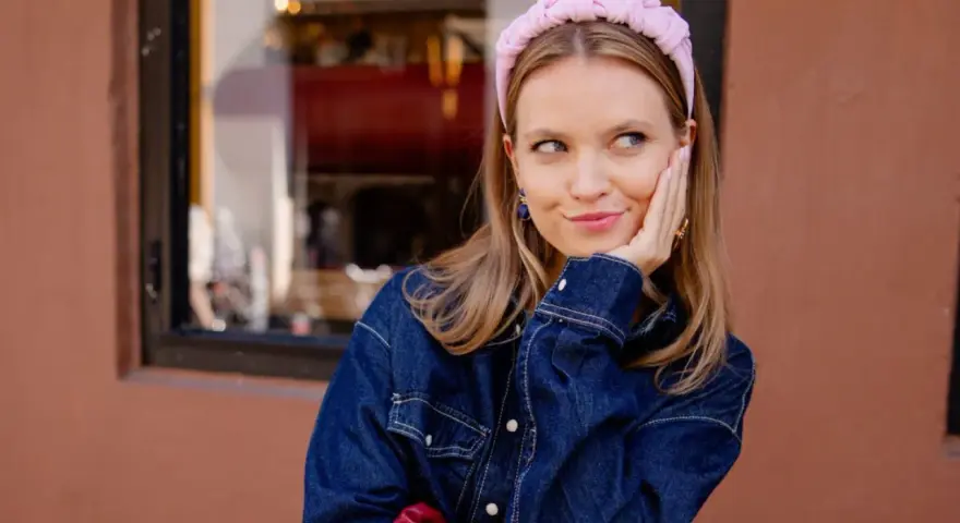 Aesthetic girl w jeansowej kurtce i różowej opasce na głowie, opiera policzek na dłoni, patrząc w bok.