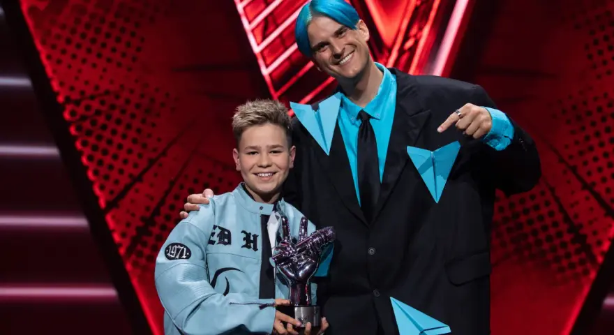 Chłopiec z błękitną kurtką i trofeum w kształcie dłoni, obok mężczyzny z niebieskimi włosami. To on wygrał The Voice Kids!
