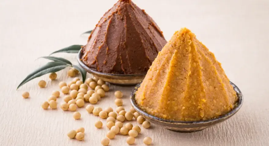 Dwie góry pasty miso, ciemna i jasna, obok ziaren soi i liści. Poznaj składniki do przygotowania tej aromatycznej pasty miso.