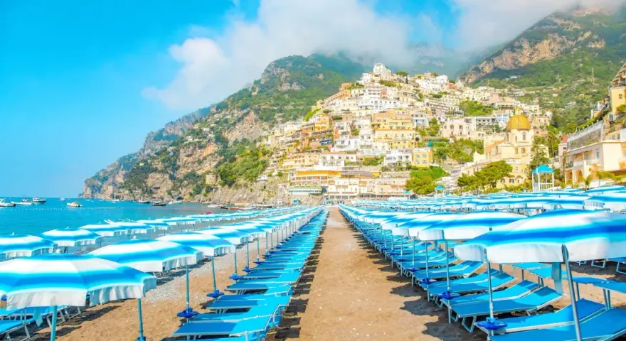 Rzędy leżaków i parasoli na plaży w Positano, Włochy. W tle widać malownicze miasteczko na zboczu góry i błękitne morze.