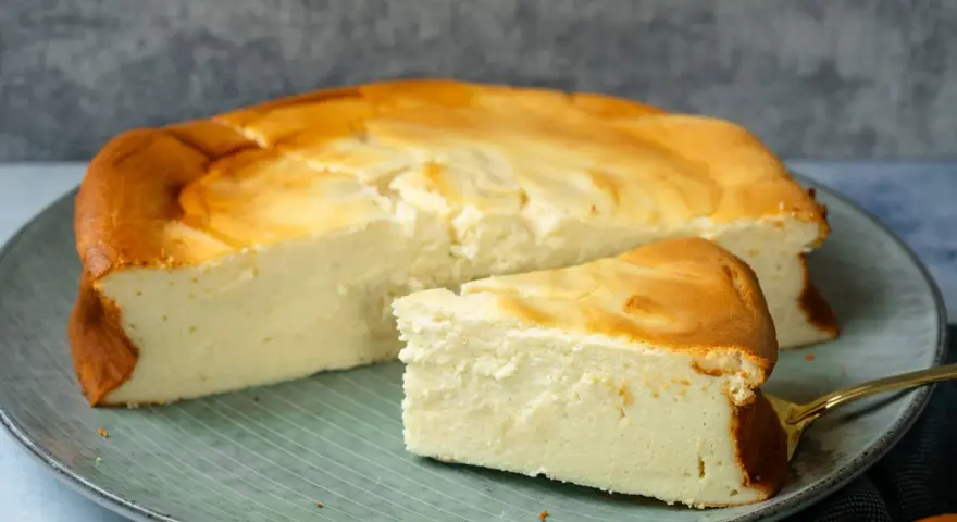 Ein saftiger Quarkkuchen ohne Boden, goldbraun gebacken, liegt auf einem Teller. Ein Stück ist bereits angeschnitten und zeigt die cremige Konsistenz.