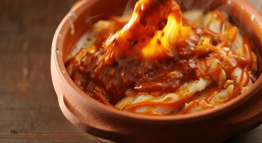 Cocochas de bacalao cubiertas con una salsa roja y burbujeante en una cazuela de barro.