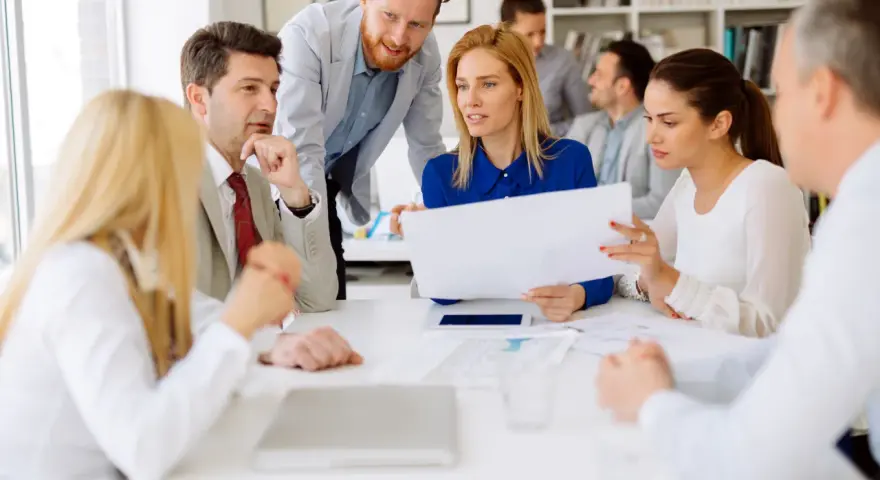 A diverse group of professionals discusses documents, possibly during board of directors recruitment or a strategic meeting.