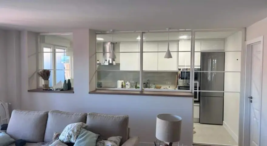Un salón moderno con sofá y lámpara, separado de una cocina blanca y luminosa por una cristalera.