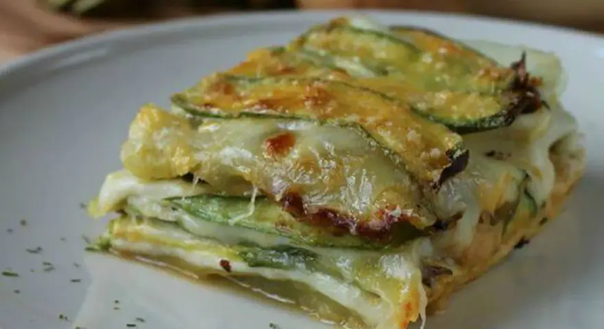 Una fetta di parmigiana di zucchine, stratificata con formaggio filante e zucchine grigliate, servita su un piatto bianco.