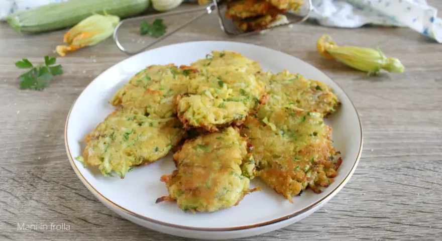 Buñuelos de calabacín dorados y crujientes en un plato blanco, con calabacines y flores en el fondo.